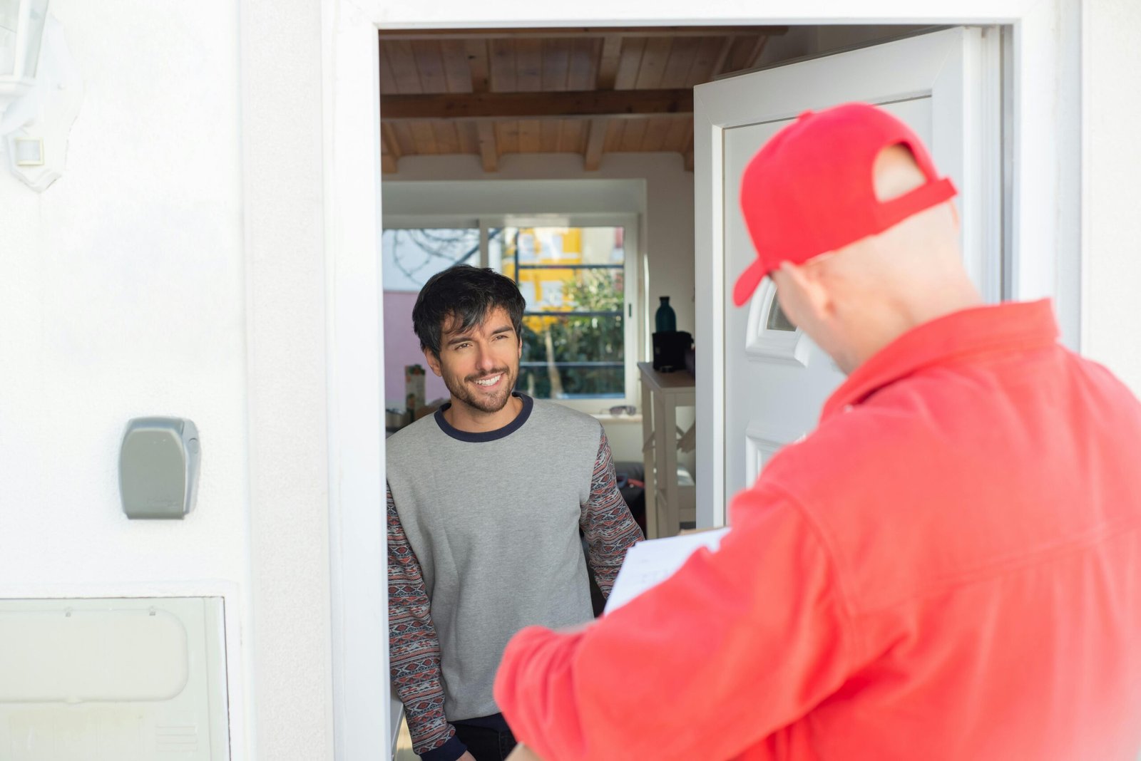 A delivery man in red uniform delivers a package to a smiling man at home.